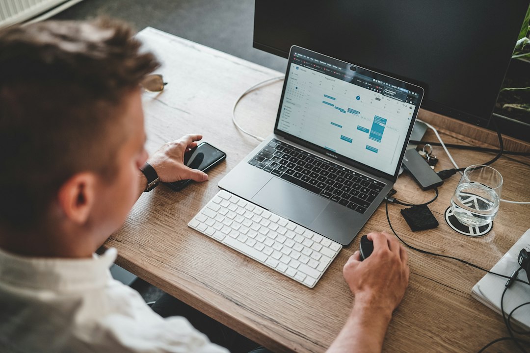 Administrator de bloc care verifică pe laptop o listă de întreținere generată automat, cu facturi de utilități și un telefon cu poza unui apometru pe birou